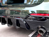A car's rear features a sporty diffuser and dual exhaust pipes, parked on an urban street with reflections of nearby buildings on its glossy black surface.