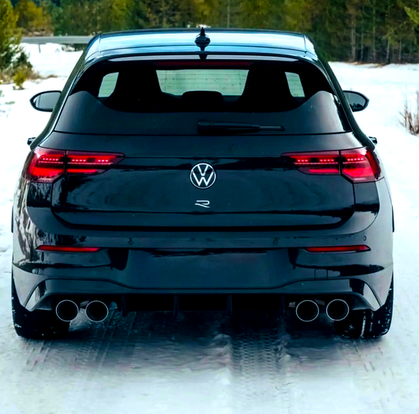 A black Volkswagen car is parked on a snow-covered road in a forested area, showing dual exhaust pipes and illuminated tail lights.