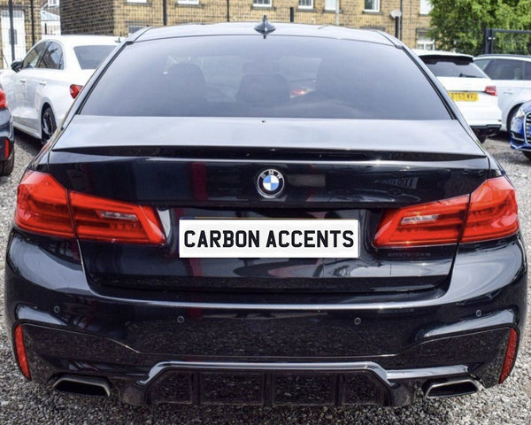 A black BMW with red tail lights is parked on gravel in a lot, surrounded by other vehicles. The license plate reads "CARBON ACCENTS."