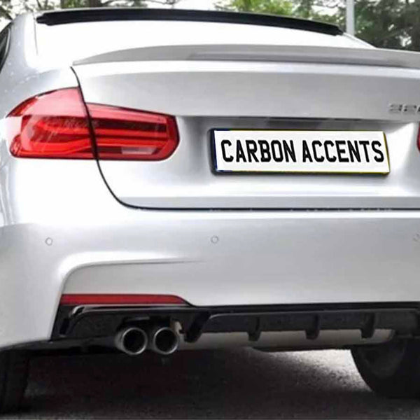A silver car is parked on a paved surface with greenery in the background. The rear features dual exhausts and a license plate reading "CARBON ACCENTS."