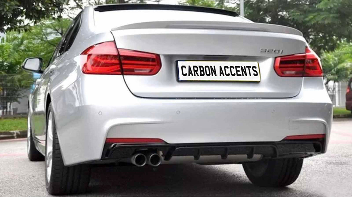 A silver car is parked on a paved surface with greenery in the background. The rear features dual exhausts and a license plate reading "CARBON ACCENTS."