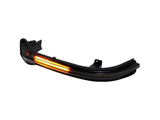 A black car side mirror indicator light emits an amber glow. It features a sleek, curved design, and is set against a plain white background.