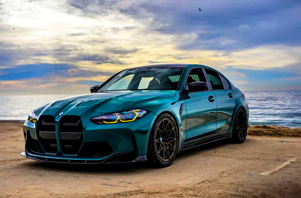 A sleek, blue sports sedan is parked beside the ocean on a sandy surface, under a dramatic, cloudy sky at sunset.