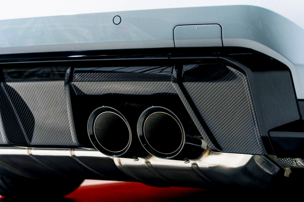 Twin exhaust pipes protruding from a vehicle's rear bumper with carbon fiber detailing, against a blurred background, highlighting the car's sporty design.