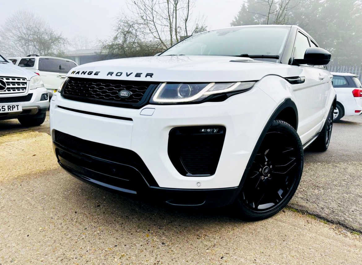 A white Range Rover parked on a concrete surface with visible branding. It is surrounded by other vehicles on a foggy day, with trees and fencing in the background.