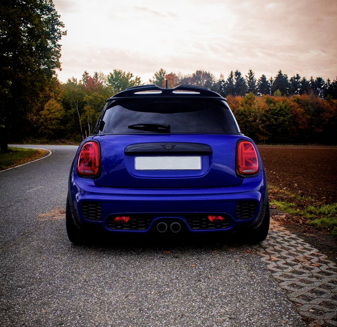 A blue compact car is parked on a winding road, surrounded by autumn trees under a cloudy sky. The car's exhaust is visible at the back.