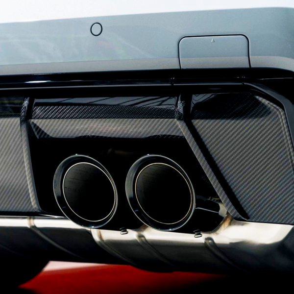 Dual exhaust pipes, surrounded by a carbon fiber diffuser, protrude from the rear of a sleek white car, emphasizing a sporty, high-performance design in an indoor setting.