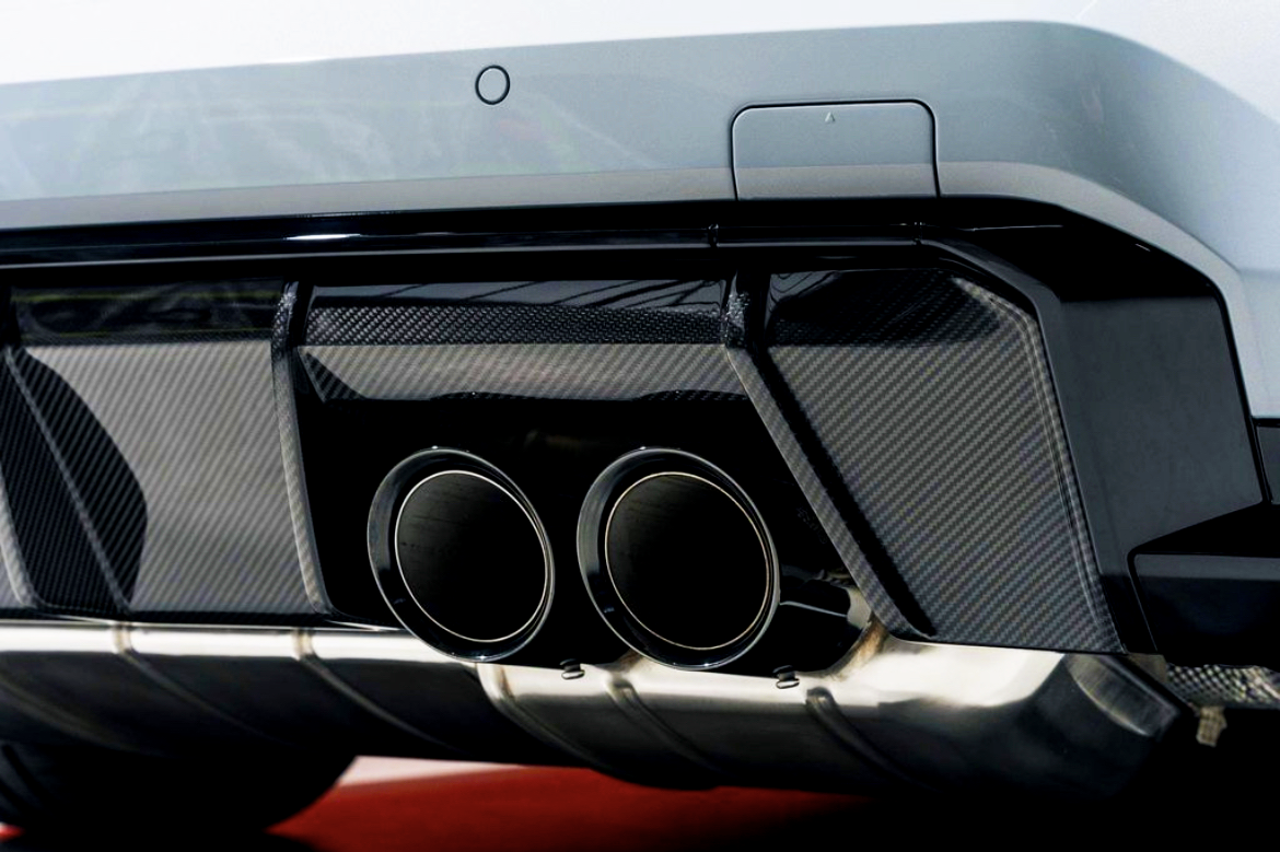 Dual exhaust pipes, surrounded by a carbon fiber diffuser, protrude from the rear of a sleek white car, emphasizing a sporty, high-performance design in an indoor setting.