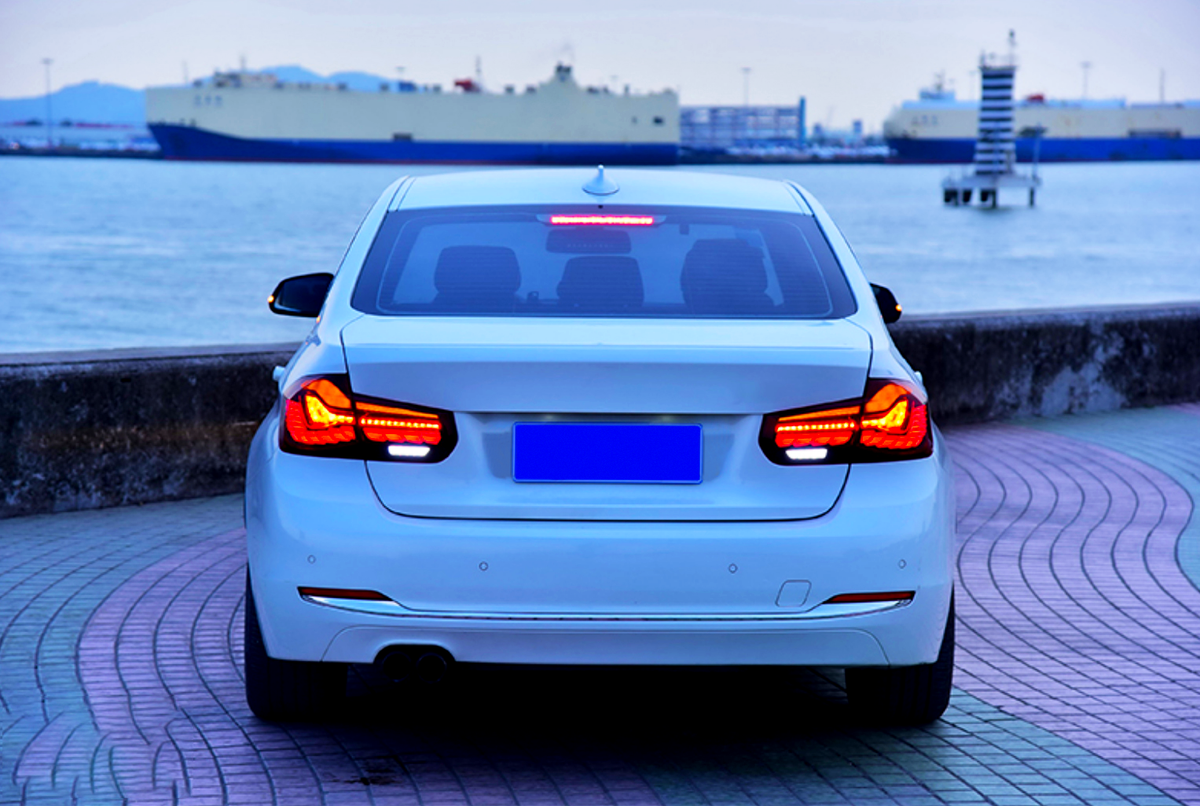 A white car is parked with illuminated taillights, facing a calm waterfront. In the background, large ships and a distant horizon are visible under a clear sky.