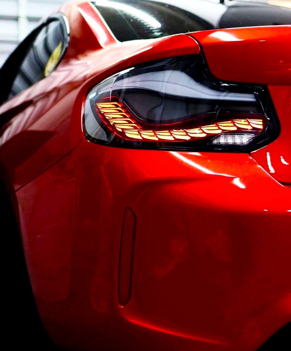 A red car's tail light, illuminated with intricate detailing, is positioned in a dimly lit garage space, reflecting light on its glossy surface.