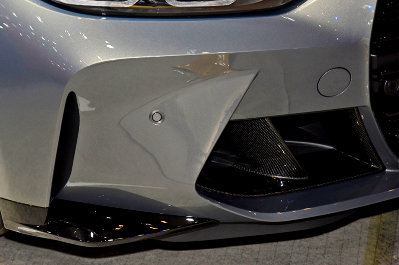 A sleek, silver car's front bumper features aerodynamic vents and textured black details, situated in a well-lit indoor setting.