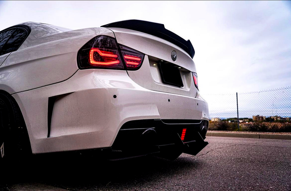 A white sports car with illuminated red taillights is parked on a paved road, near a fence, under a cloudy sky.