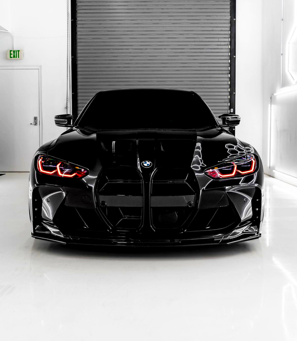 A sleek black BMW with illuminated red headlights is parked in a white room in front of a metal shutter door. An "EXIT" sign is visible on the left.