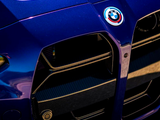 A blue car's front grille features a prominent BMW emblem, showcasing sleek design elements and curves. The sunlight highlights the vehicle's glossy finish on a clear day.