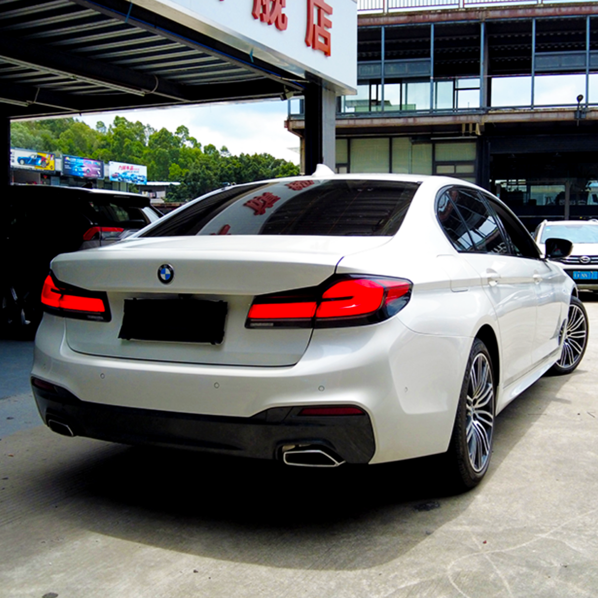 A white BMW parked under a structure with a partly visible Chinese sign, surrounded by other cars and greenery in the background.