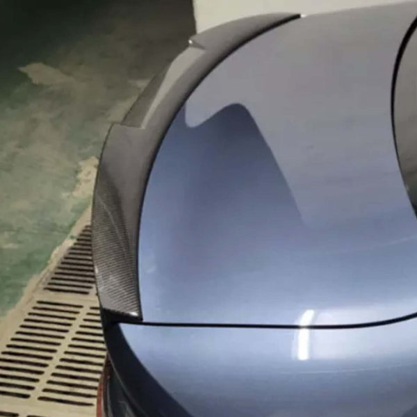 A car trunk with a carbon fiber spoiler is parked in an indoor garage with a green and beige floor, next to a drainage grate.