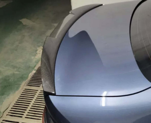 A car trunk with a carbon fiber spoiler is parked in an indoor garage with a green and beige floor, next to a drainage grate.