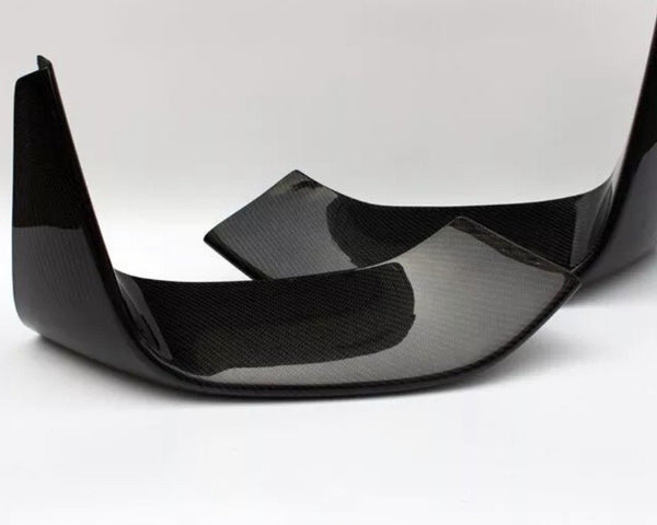 Two carbon fiber car parts, possibly rear exhaust diffusers, are positioned diagonally on a solid white surface, highlighting their glossy black finish and textured pattern.