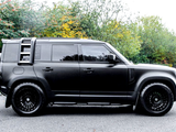 A black SUV is parked on a road, featuring black wheels and tinted windows, with a lush, green, and slightly autumnal forest backdrop.
