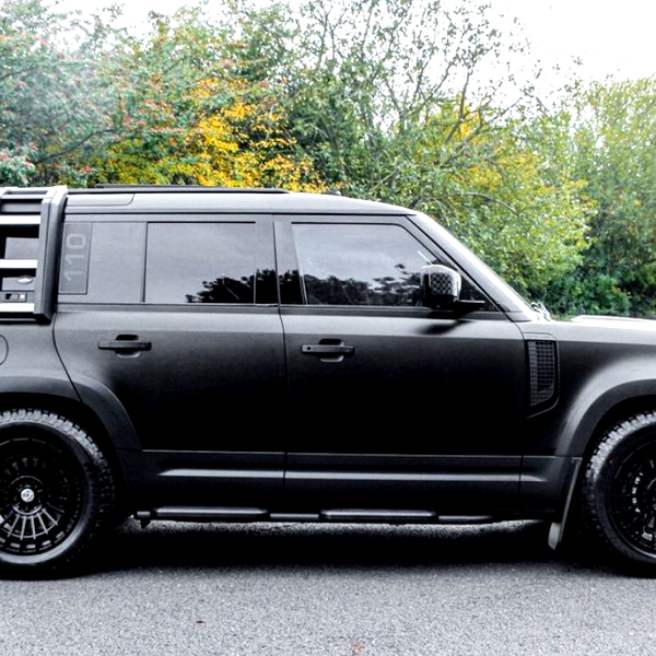 A black SUV is parked on a road, featuring black wheels and tinted windows, with a lush, green, and slightly autumnal forest backdrop.
