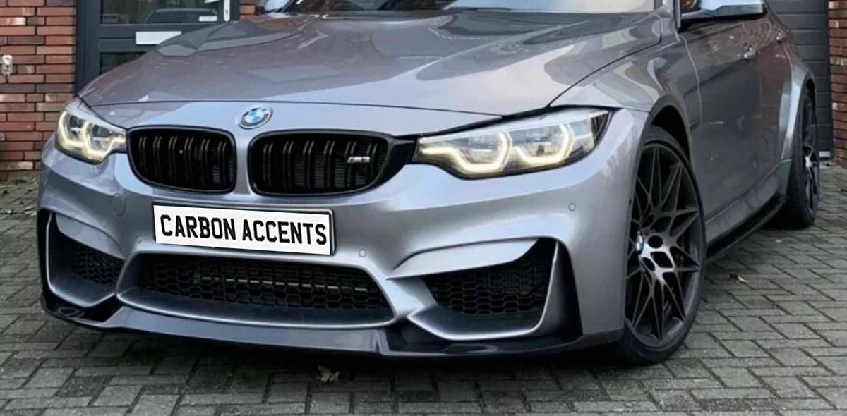 A sleek, grey sports car is parked on a brick pavement, displaying a license plate that reads "CARBON ACCENTS." The headlights are illuminated, and the front grille is prominent.