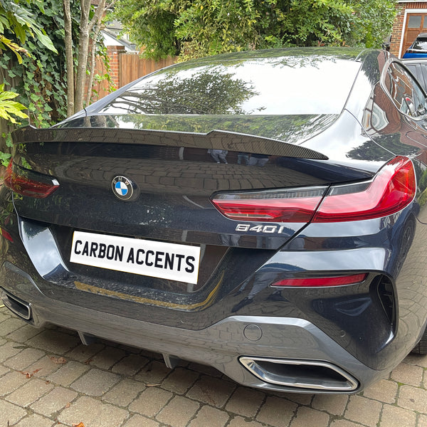 A black BMW 840i is parked on a stone driveway surrounded by greenery. The license plate reads "CARBON ACCENTS."