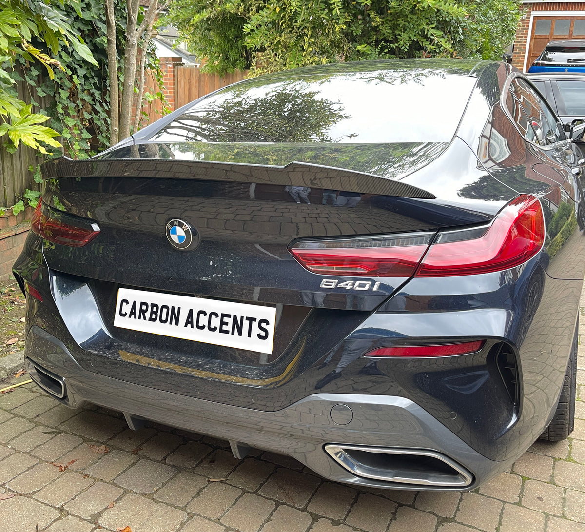 A black BMW 840i is parked on a stone driveway surrounded by greenery. The license plate reads "CARBON ACCENTS."