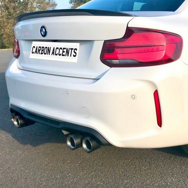A white BMW with dual exhausts, parked on a sunny road. The license plate reads "CARBON ACCENTS." Red taillight and carbon fiber spoiler enhance the sporty look.