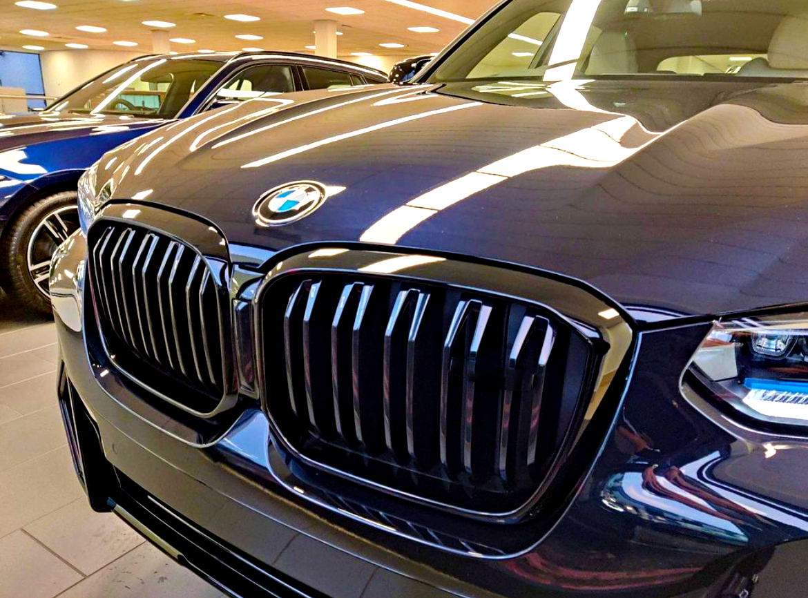 The front of a dark-colored car with a prominent grille and BMW logo is parked indoors alongside other vehicles under bright ceiling lights.