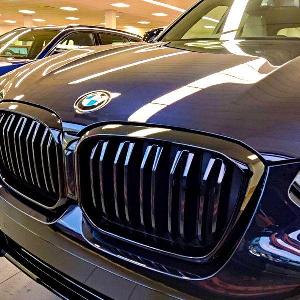 The front of a dark-colored car with a prominent grille and BMW logo is parked indoors alongside other vehicles under bright ceiling lights.
