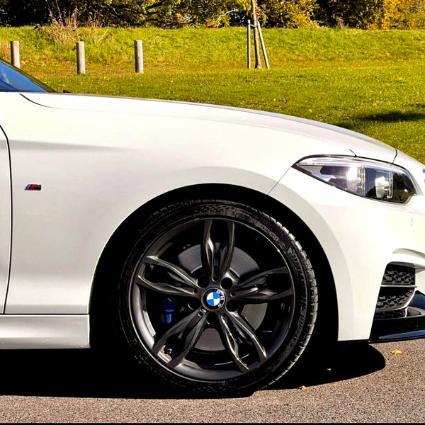 A white car is parked on a paved road. Its front wheel is turned slightly, showcasing a five-spoke alloy rim. The background features a grassy hill and wooden posts.