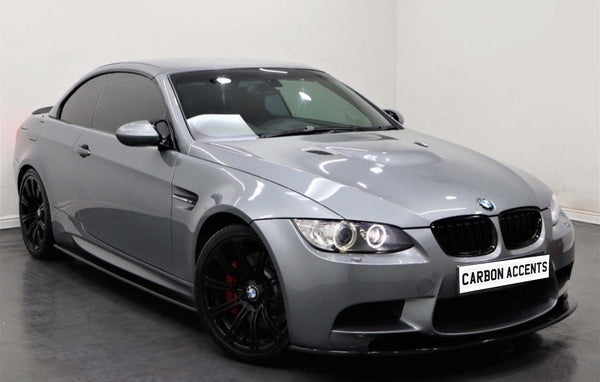 A sleek, silver sports car with tinted windows and black rims sits in a white showroom. The license plate reads "CARBON ACCENTS."