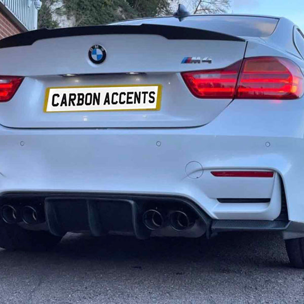 A white BMW M4 displays illuminated red taillights. The license plate reads "CARBON ACCENTS." The car is parked near a building with red brick and a gray door.