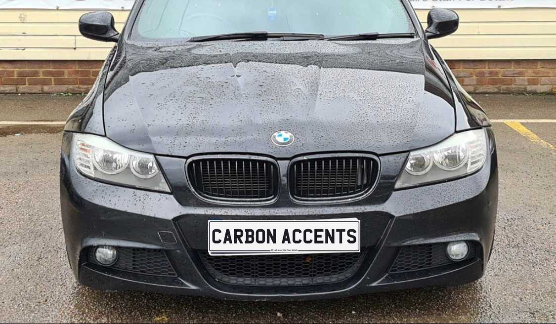 A black BMW car with water droplets on the hood is parked. The license plate reads "CARBON ACCENTS." The background features a brick wall and pavement.