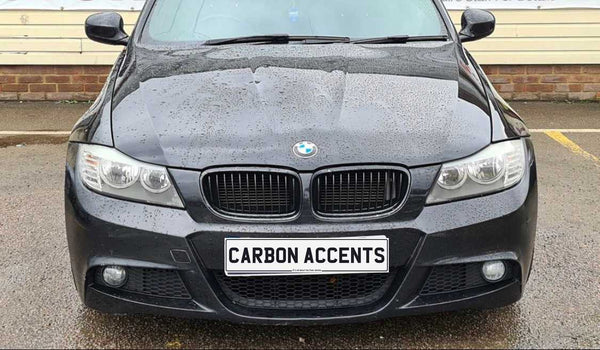 A black BMW car with water droplets on the hood is parked. The license plate reads "CARBON ACCENTS." The background features a brick wall and pavement.