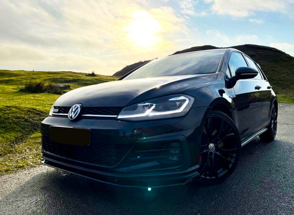 A black Volkswagen car, parked on a paved road, is illuminated by a setting sun, with grassy hills in the background. Text visible: "GTI" on the front grille.