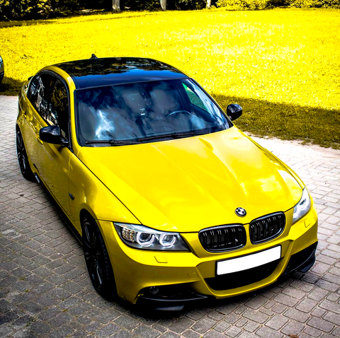 A yellow car is parked on a paved driveway. It features a glossy black roof and is surrounded by a grassy area with scattered trees in the background.