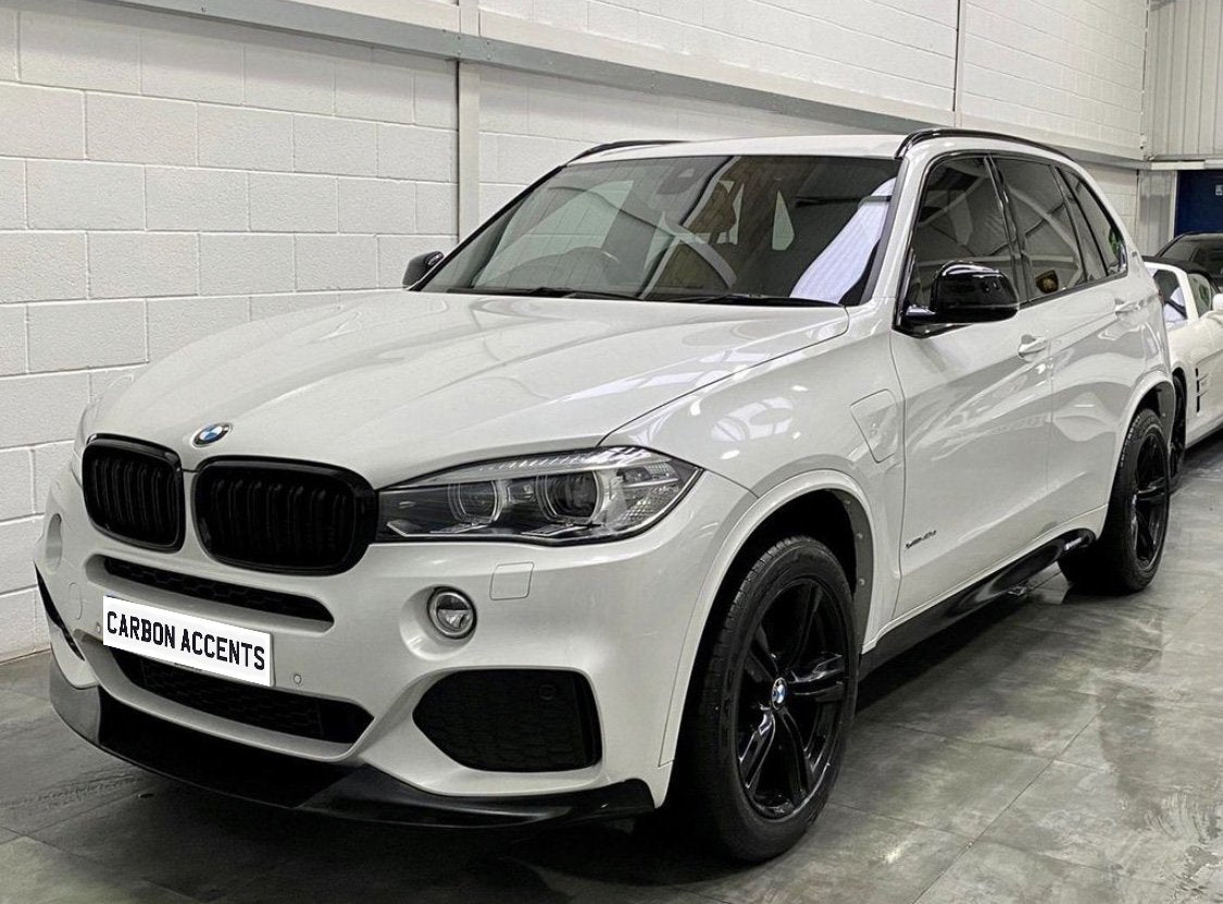 A white BMW SUV is parked indoors on a concrete floor, featuring black wheels and a front license plate that reads "CARBON ACCENTS."