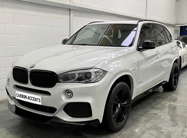 A white BMW SUV is parked indoors on a concrete floor, featuring black wheels and a front license plate that reads "CARBON ACCENTS."
