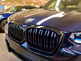 A shiny BMW car sports a prominent grille in a well-lit showroom, surrounded by other vehicles and illuminated by overhead lights.
