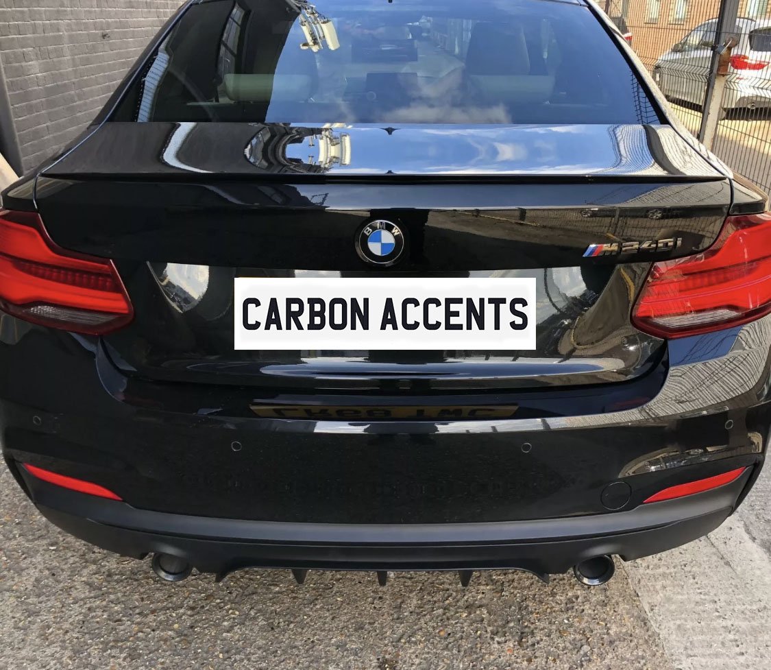 A black BMW car is parked in an outdoor setting, displaying a "M240i" badge. Text reads "CARBON ACCENTS." The background includes a brick building and a fenced area.
