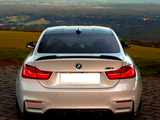 A silver BMW M4 is parked on a cobblestone path, rear view facing the camera, with a scenic backdrop of green fields and distant mountains under a clear sky.