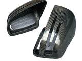 Two carbon fiber car mirror covers lie on a gray fabric surface, each featuring sleek, vent-like slits for aerodynamic styling.