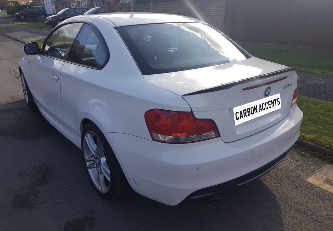 A white BMW 123d, parked on a street, displays a carbon spoiler. Text on the rear reads "CARBON ACCENTS." Other cars and houses are visible in the background.