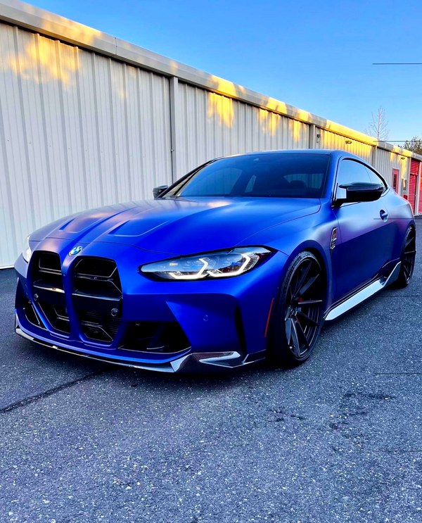 A blue sports car is parked on a paved area, with a large building featuring corrugated metal walls in the background under a clear blue sky.