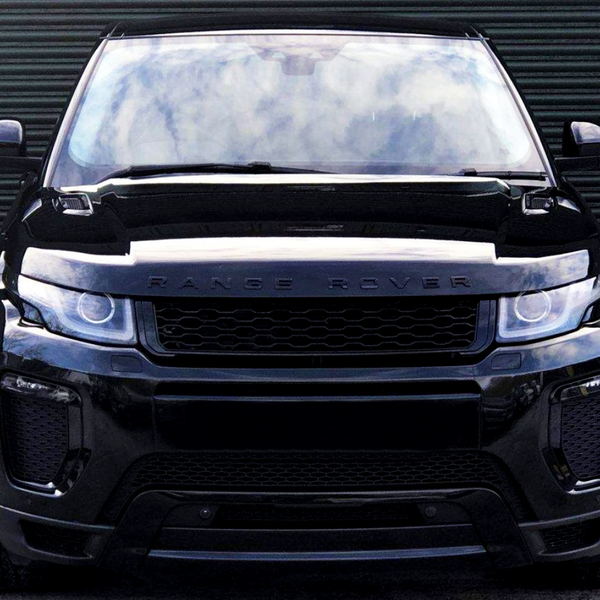 A black Range Rover stands stationary, facing forward, on concrete ground against a closed, corrugated metal door. The vehicle's headlights and grill are prominent. Text on the hood: "RANGE ROVER."