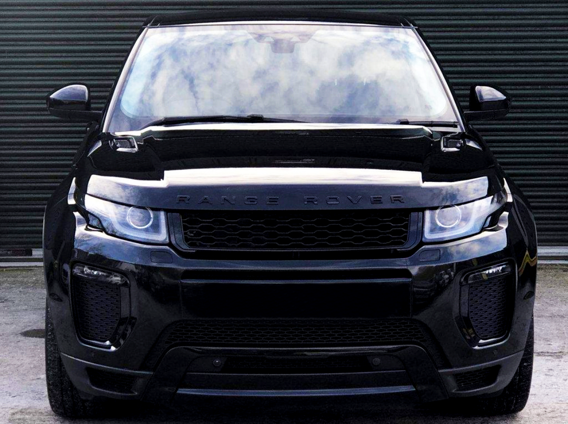 A black Range Rover stands stationary, facing forward, on concrete ground against a closed, corrugated metal door. The vehicle's headlights and grill are prominent. Text on the hood: "RANGE ROVER."
