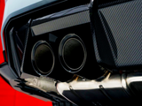 Twin exhaust pipes, metallic and polished, positioned at the rear of a vehicle with carbon fiber trim, against a bright red backdrop, highlighting the sleek automotive design.