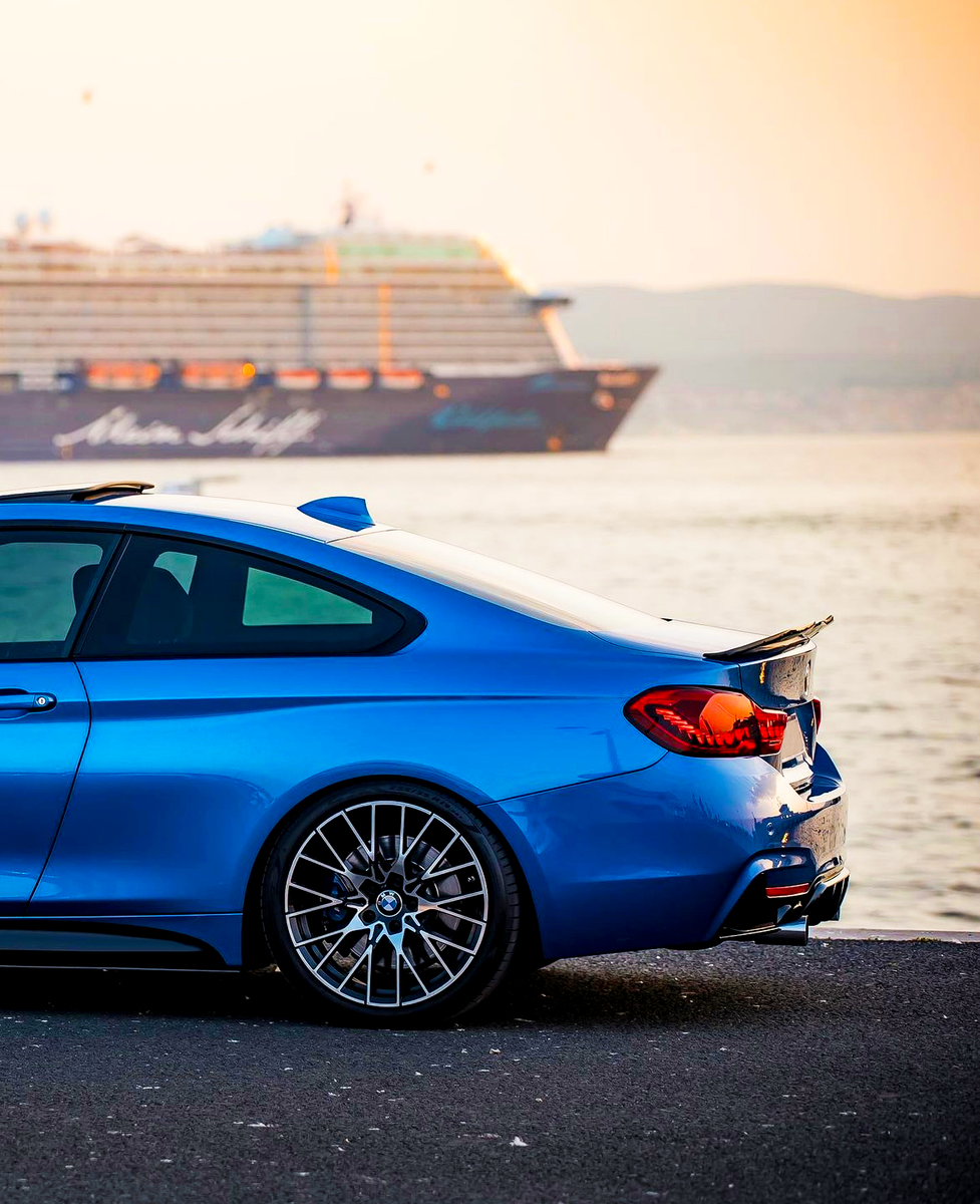 A blue sports car is parked by a waterfront at sunset, with a large cruise ship labeled "Mein Schiff" passing by in the background.