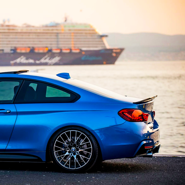 A blue sports car is parked by a waterfront at sunset, with a large cruise ship labeled "Mein Schiff" passing by in the background.